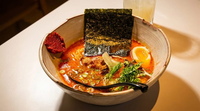Spicy ramen bowl with noodles, soft-boiled egg, nori, and chili paste on a white table.