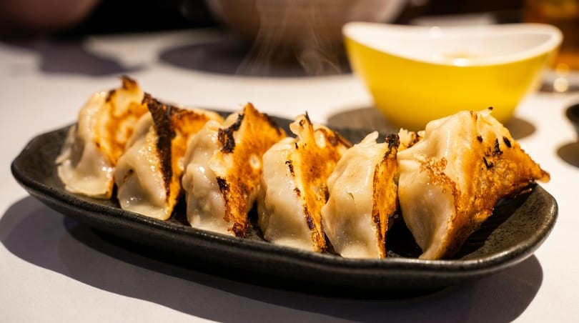 Steaming pan-fried gyoza with crispy golden-brown bottoms on a dark plate, with a yellow bowl.