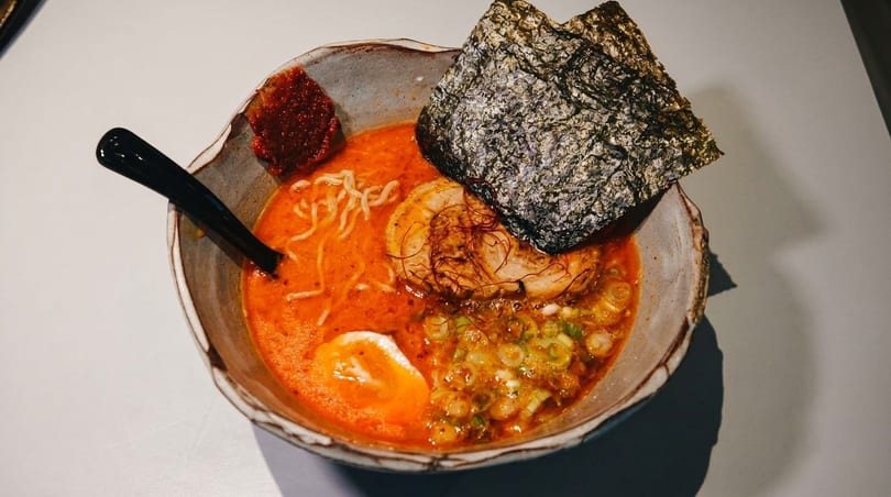 A vibrant bowl of spicy ramen with pork, egg, nori, noodles, and green onions.