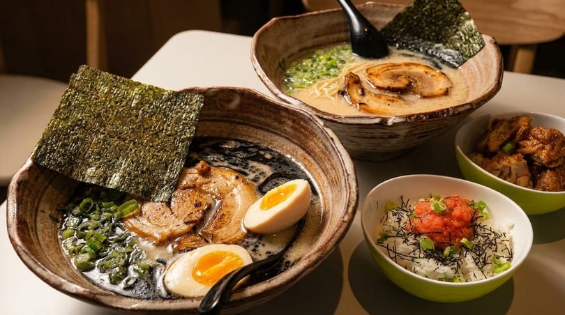 Two bowls of ramen with pork, egg, and nori, alongside rice and fried chicken side dishes.