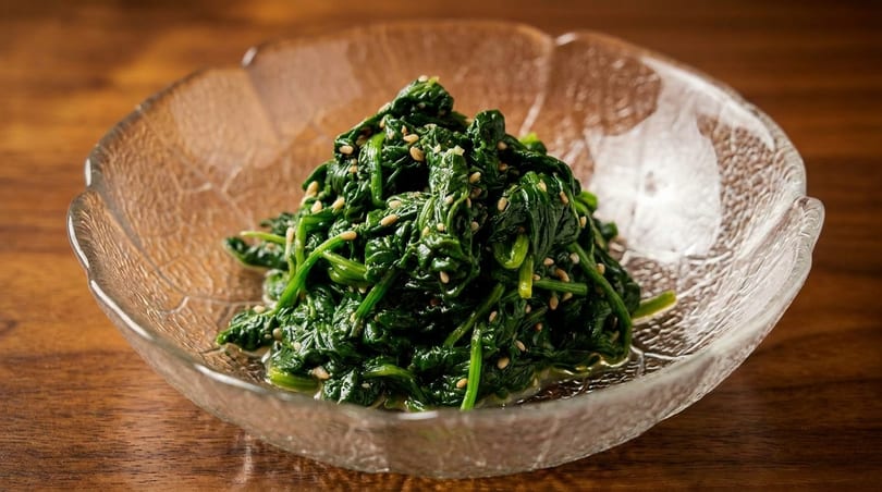 Cooked spinach topped with sesame seeds in a clear glass bowl on a wooden surface.