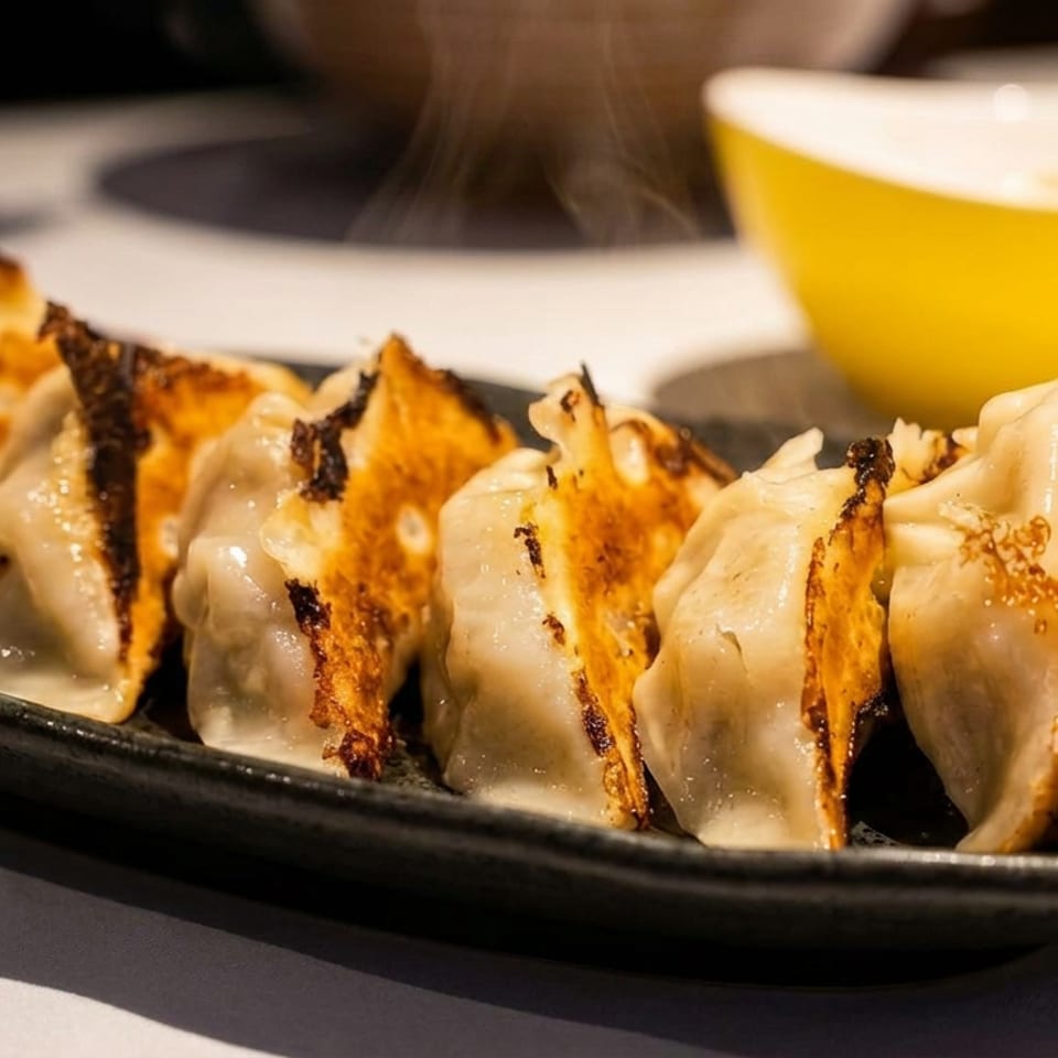 Golden-brown pan-fried dumplings arranged on a black plate, with a yellow bowl and dipping sauce in the background.